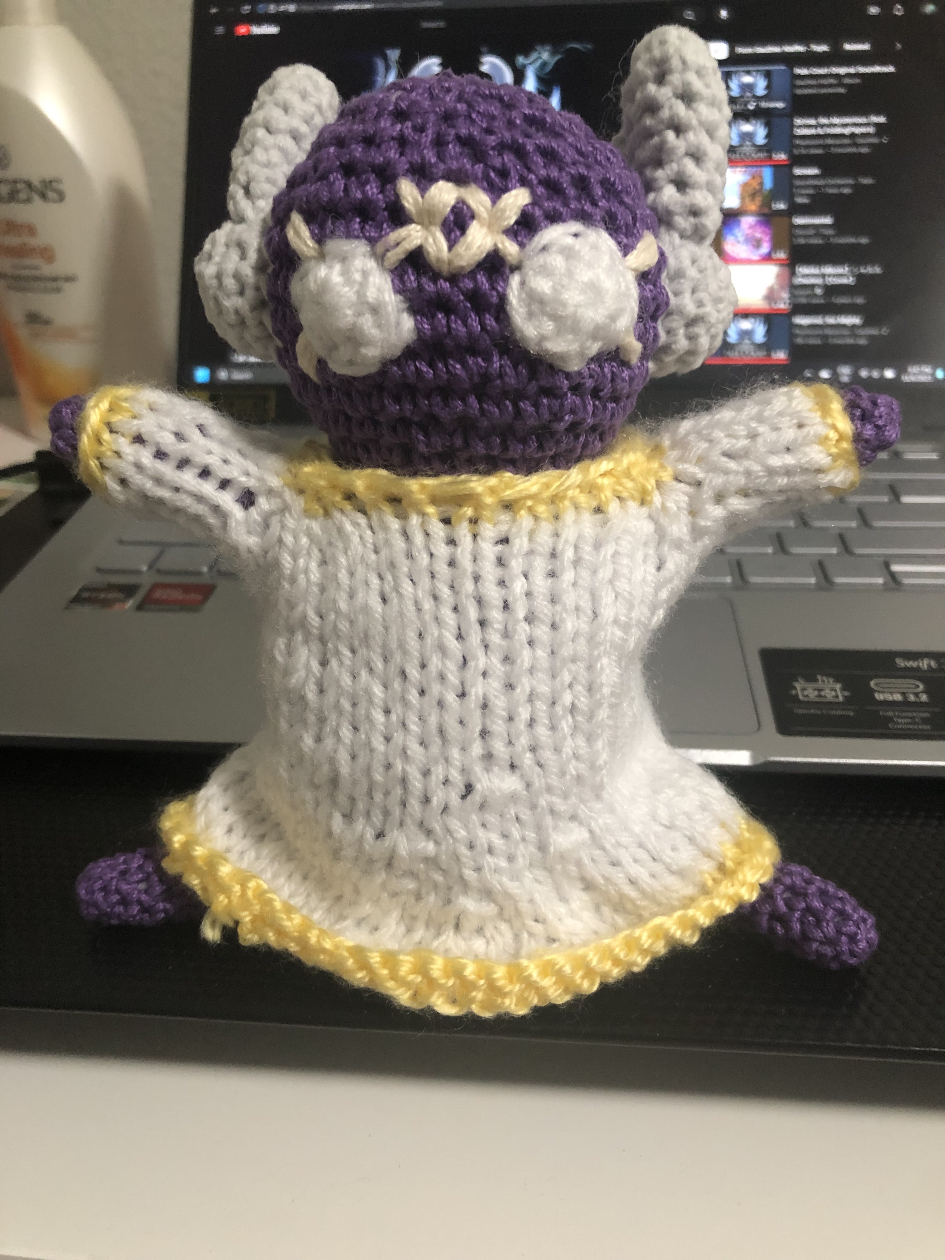 My crocheted doll of Unparalleled Innocence. They're purple and have white eyes. They have two cream markings radiating outwards from each of their eyes, and a small cream diamond shaped marking on their forehead that has similar radiating markings on the two horizontal corners. Their cloak is white with yellow hems. Their antennae are lobe shaped.